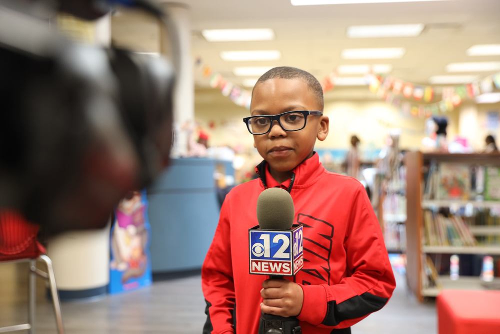 A young male student is holding a microphone while standing in front of a camera inside Sherman Avenue's library.