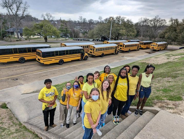 14 Vicksburg Junior High Students wearing yellow shirts stand on the steps in front of school buses