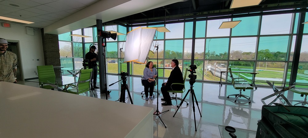 Muriel Summers and Superintendent Shealy are seated in front of several cameras and lighting during an interview. Film crew members are in the background of a room with green and clear windows overlooking the football stadium at Vicksburg High School 