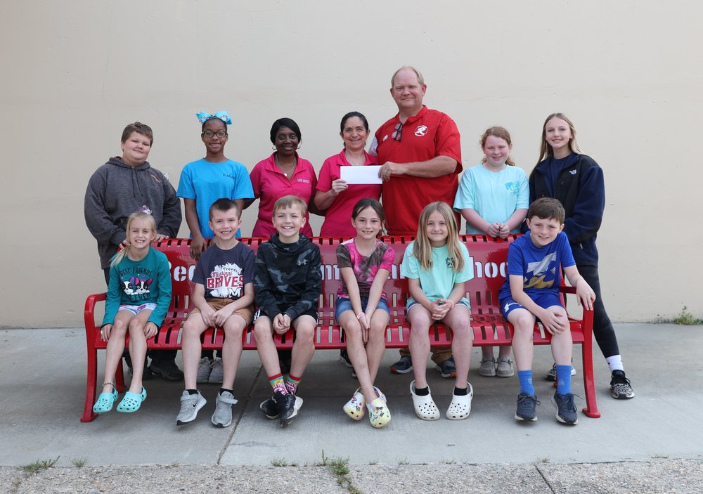 Six elementary students are sitting on a red bench while four other students are standing behind with Redwood's principal and two female representatives from United Way
