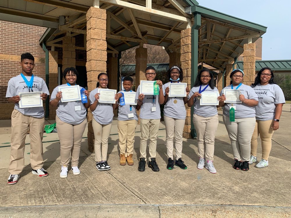 9 African American students wearing blue shirts and khaki pants hold up certificates and ribbons
