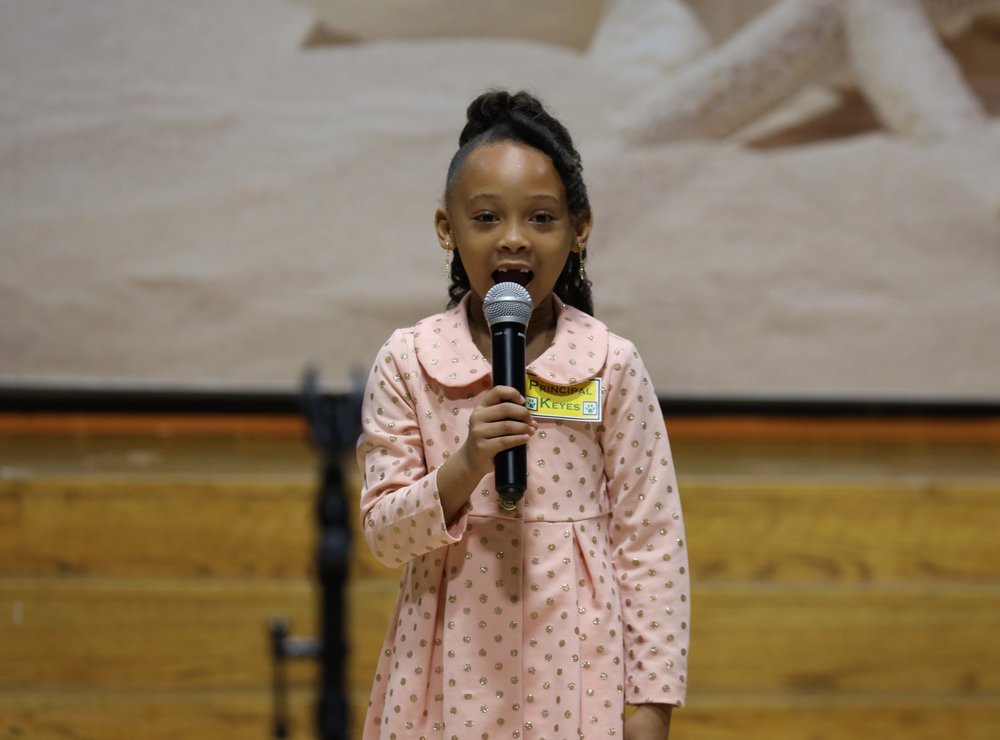 A young female student speaking into a microphone wearing a pink dress.