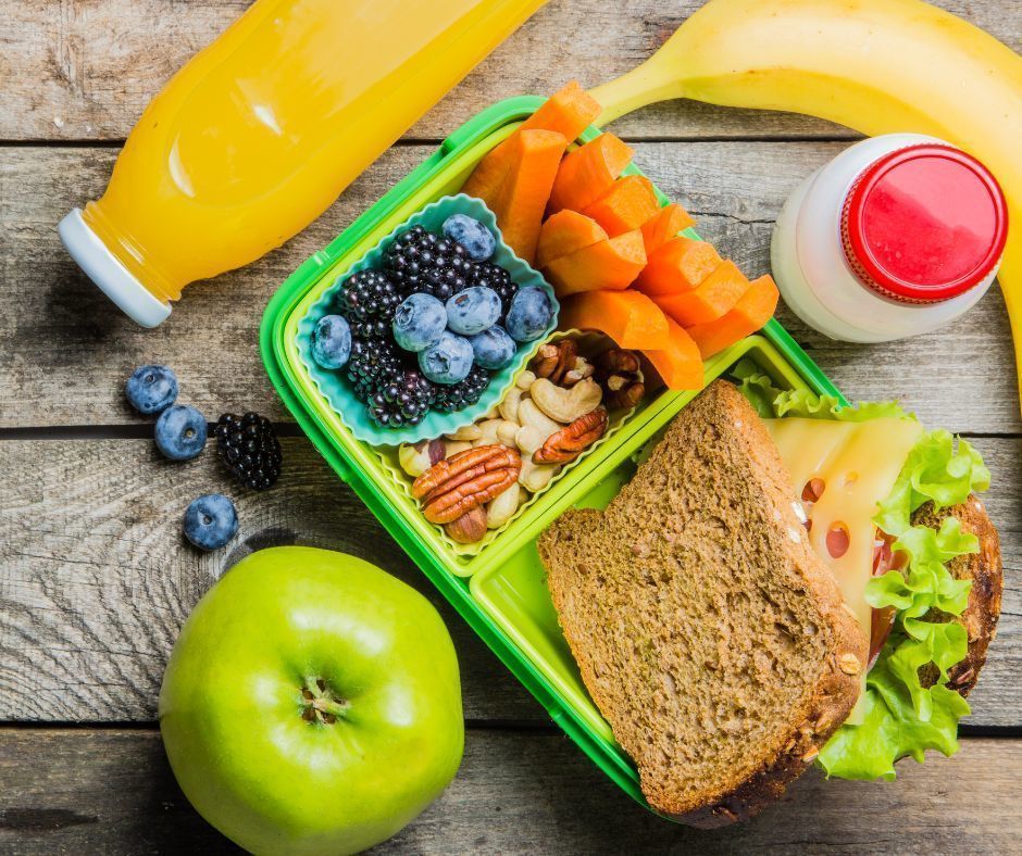 A green lunchbox filled with brightly colored food and snacks sitting on a picnic table.