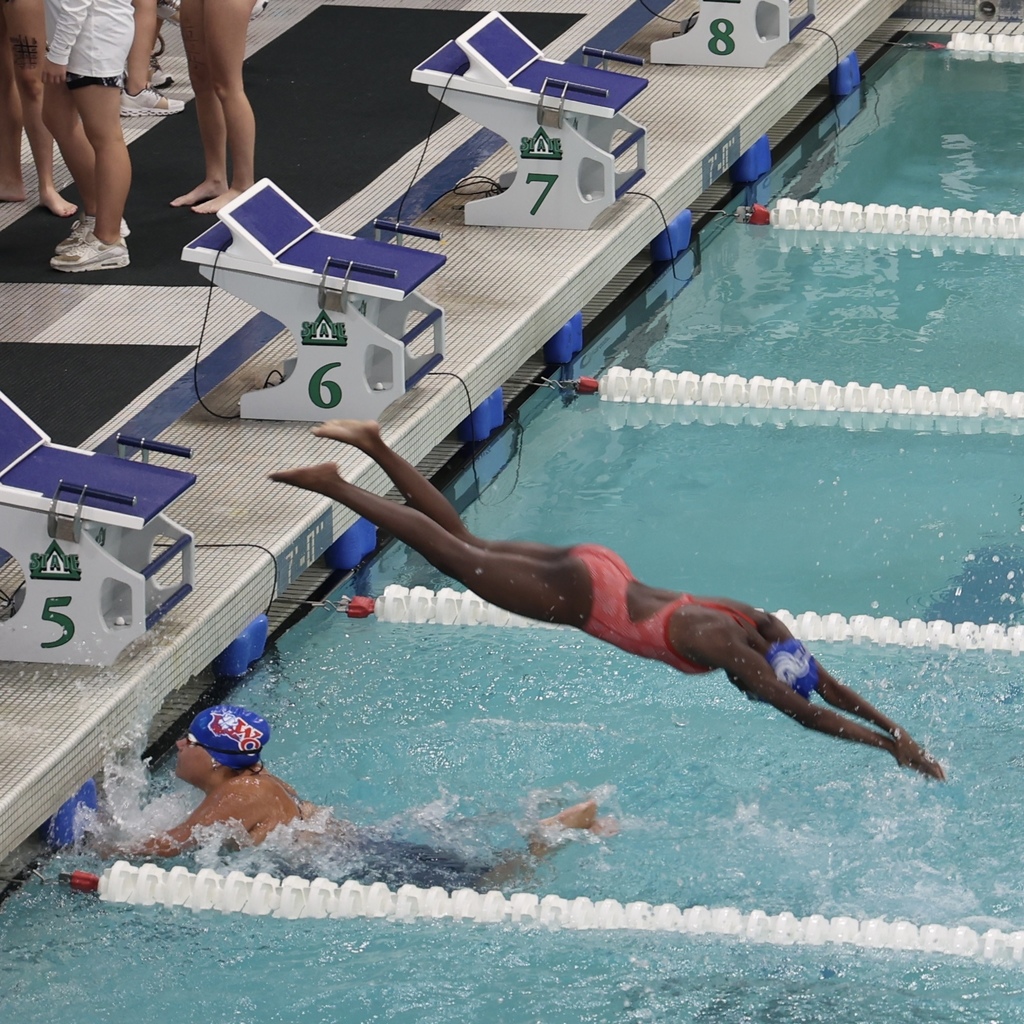 black female swimmer wearing red bathing suit and blue swim cap dives into a pool during a relay race