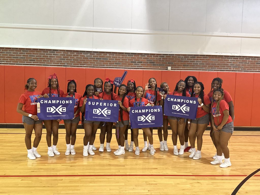 A group of sixteen cheerleaders holding purple signs in a gymnasium.