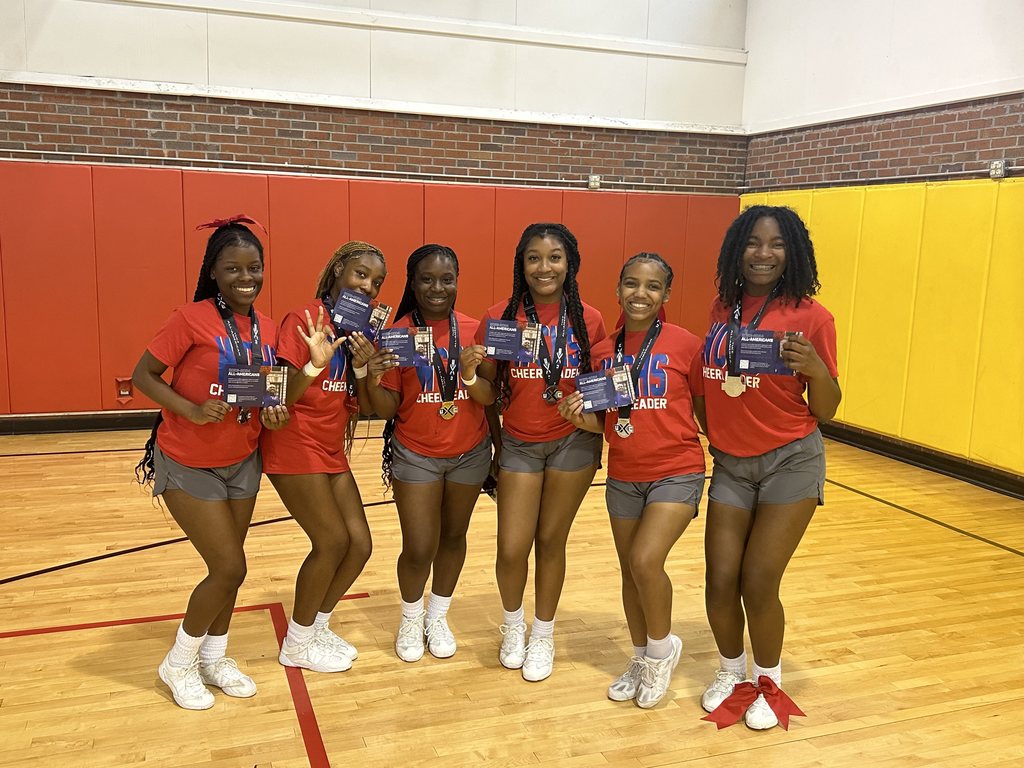 Six cheerleaders pose for a photo with their awards inside a gymnasium.